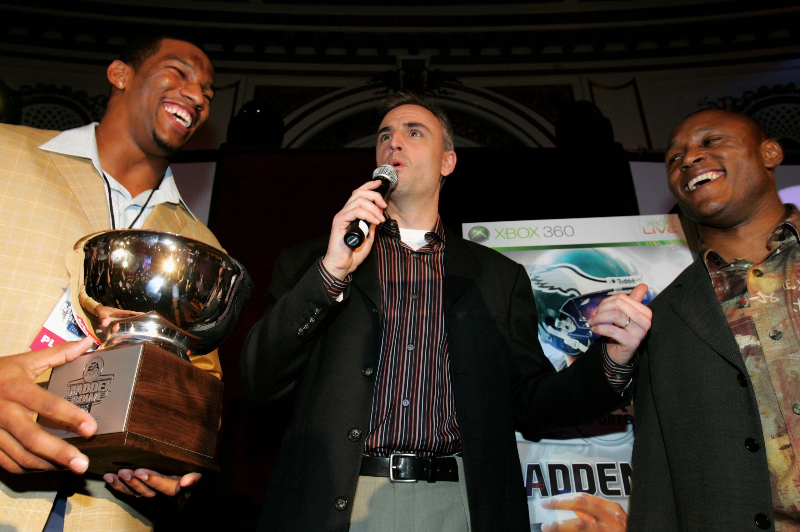 DETROIT, MICHIGAN - FEBRUARY 2:  Alex Smith of the Tampa Bay Buccaneers is presented with the trophy by Barry Sanders as announcer Trey Wingo looks on after Smith defeated Mark Clayton of the Baltimore Ravens to win the EA Sports Madden Bowl, on February