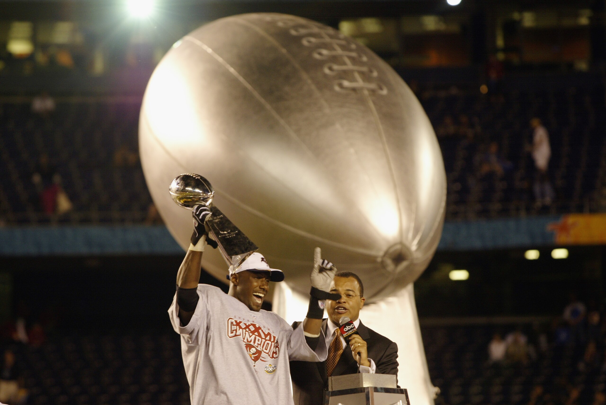 SAN DIEGO - JANUARY 26:  Super Bowl MVP Dexter Jackson #34 of the Tampa Bay Buccaneers celebrates on the victory stage as he holds the Vince Lombardi Trophy while wearing his Super Bowl Champions tee shirt and cap as ABC Sports commentator Mike Tirico loo