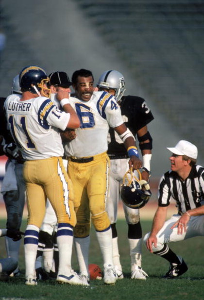 LOS ANGELES - DECEMBER 18:  Quarterback Ed Luther #11 of the San Diego Chargers tucks the shoulder pads into the jersey of teammate Chuck Muncie #46 during a game against the Los Angeles Raiders at the Los Angeles Memorial Coliseum on December 18, 1983 in